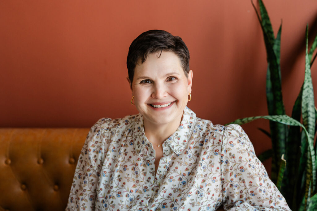 A caucasian woman smiles in front of an orange backdrop at Sol and Sage studios in Richardson, TX. She's wearing a floral shirt.