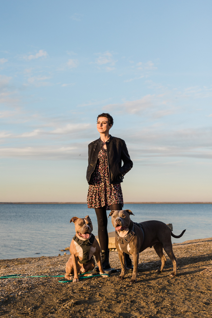Woman standing by a lake lit by sunlight with her two dogs.