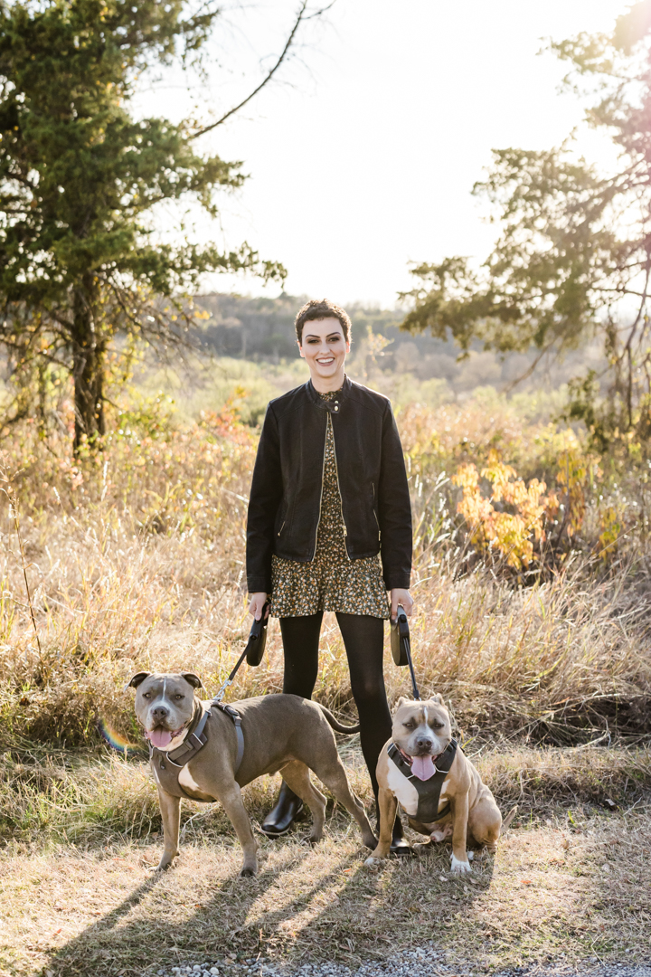 Dallas Brand Photographer; photo of a woman smiling and holding the leashes of her two pit bull dogs in front of an outdoor, nature setting