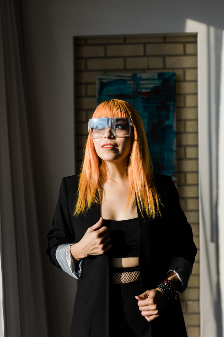Personal Brand Dallas Fort Worth; a woman with dyed orange hair, futuristic glasses, and wearing all black looking mysteriously off to the side in front of a brick background