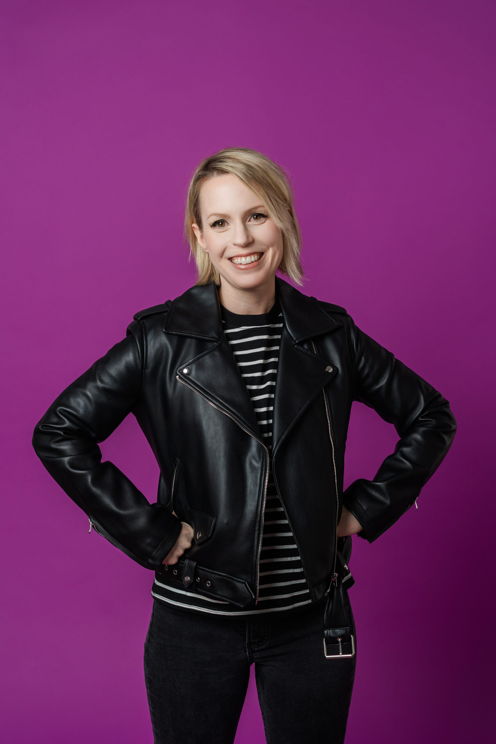 a woman wearing a black leather jacket and striped T-shirt with her hands on her hips standing in front of a purple backdrop