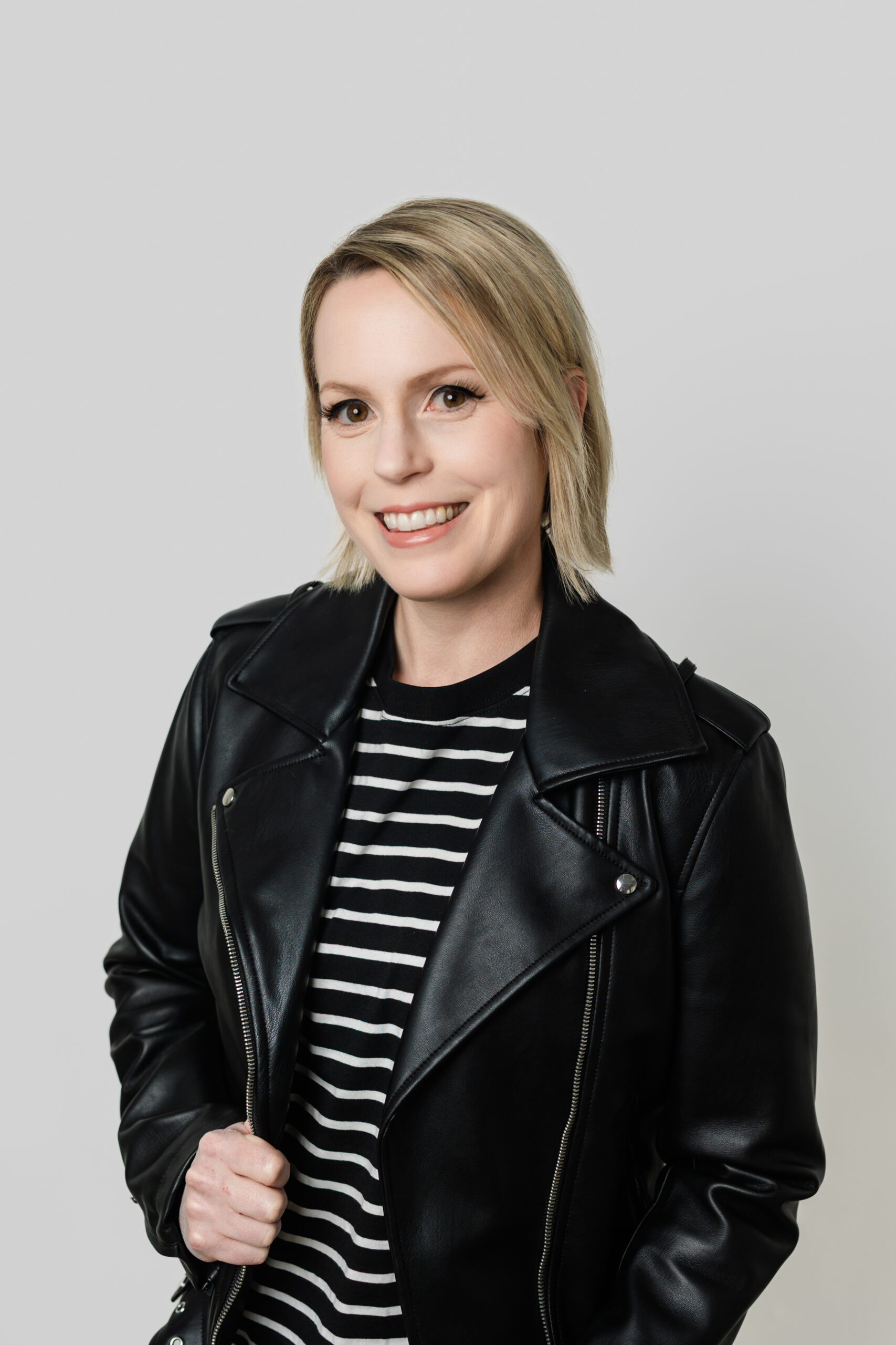 a woman wearing a black leather jacket taking a headshot in front of a gray wall 