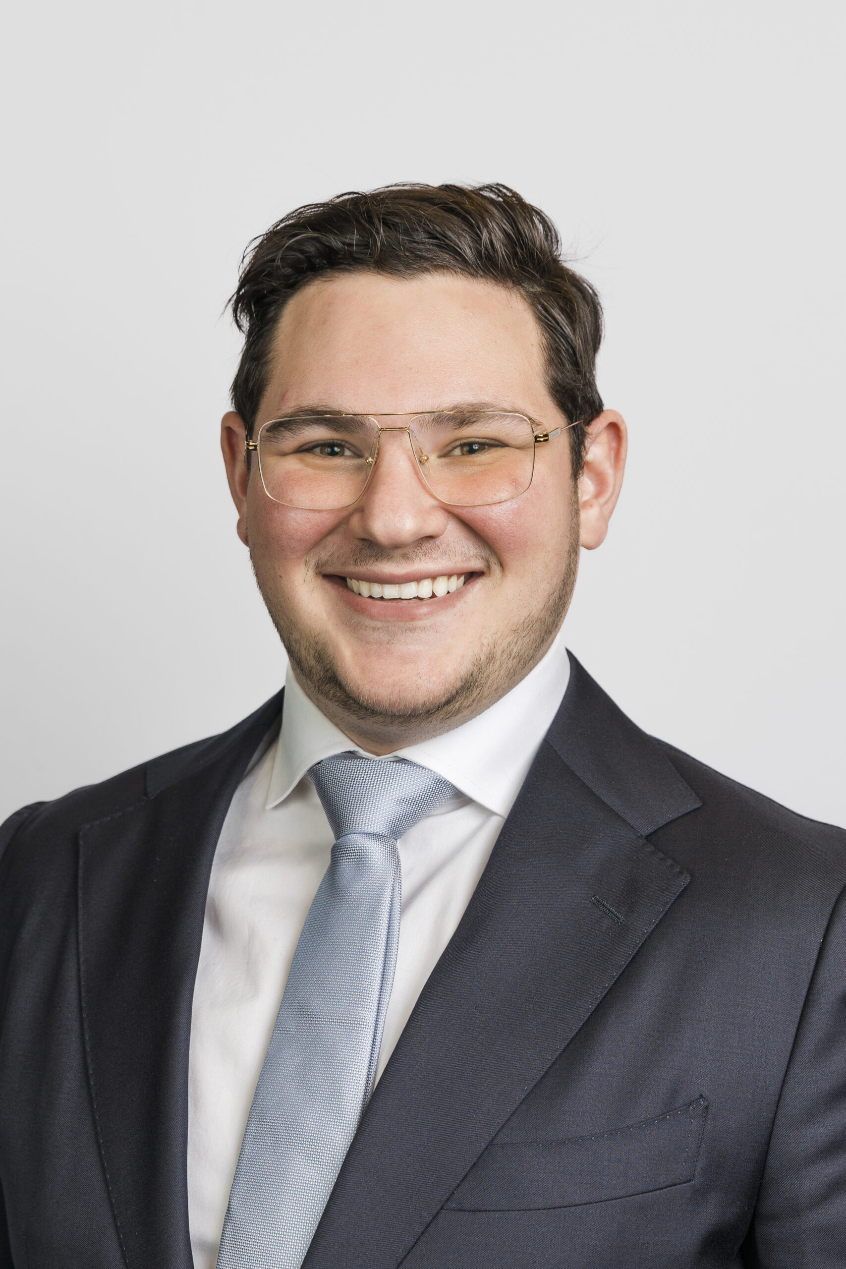 a man in a blue tie, white button up, and suit jacket wearing glasses smiling and taking a corporate headshot 