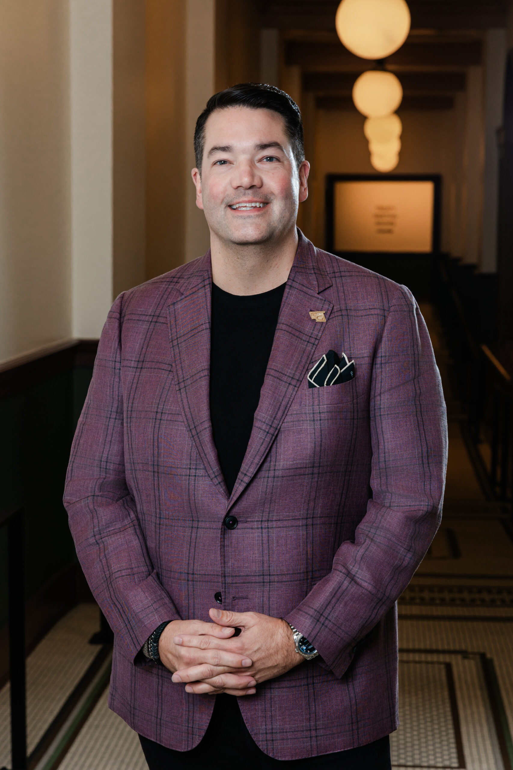a man wearing a black shirt and purple blazer taking a corporate headshot he is holding his hands crossed in front of him 