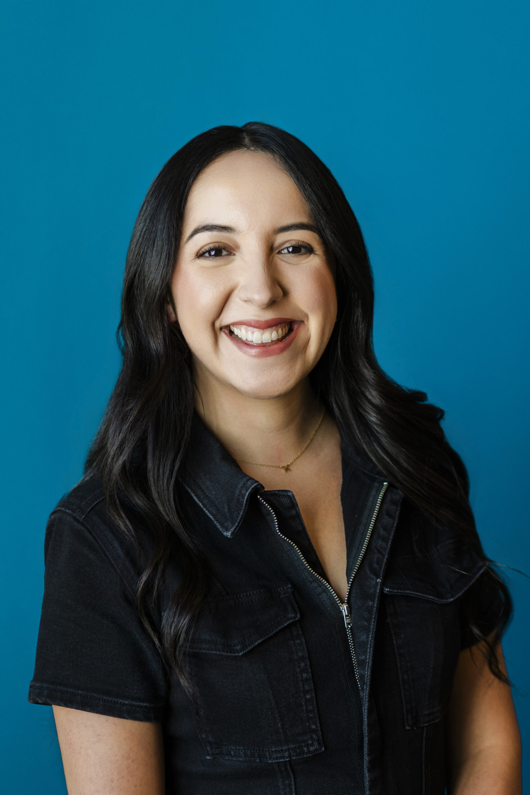 a woman wearing a black zip up taking a headshot in fronto f a blue wall 